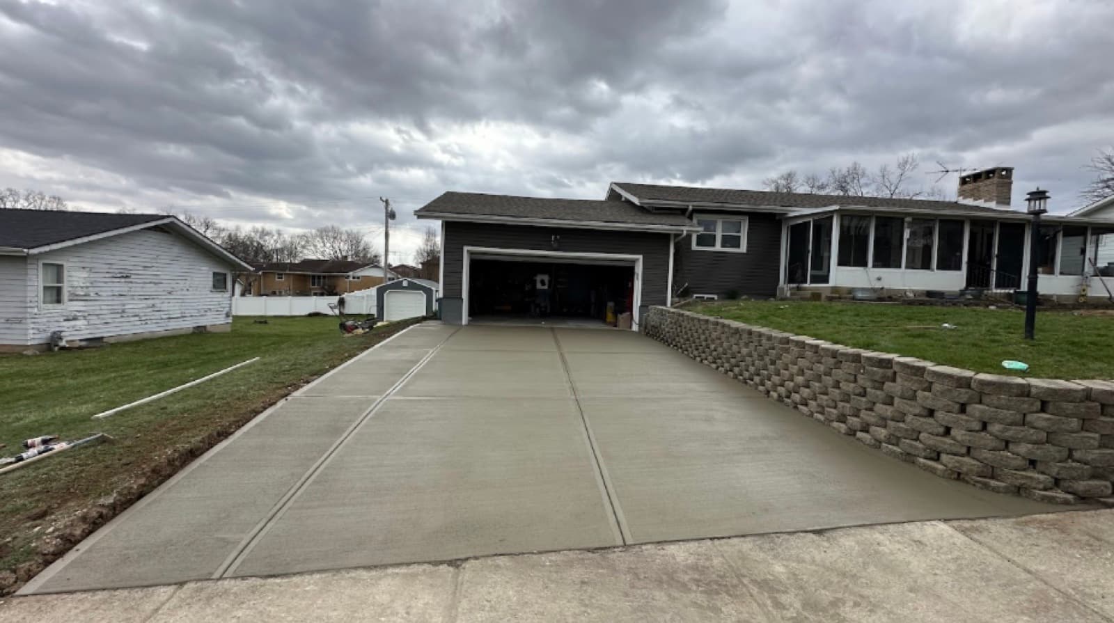 Long concrete driveway leading to a ranch home with stone retaining wall under cloudy sky